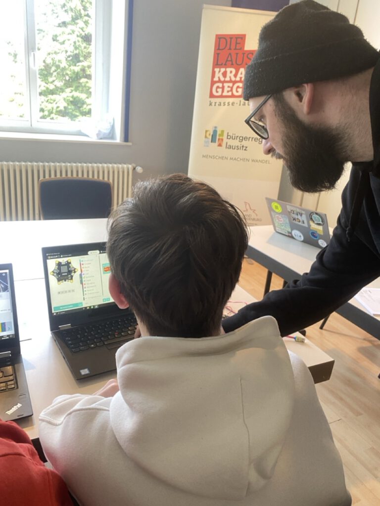 Ein Kind programmiert an einem Computer und Max Paulus erklärt gerade die aktuellen Schritte.